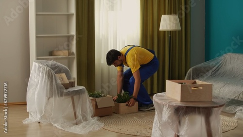 Close up shot of apartment living room. Delivery service worker in uniform brings boxes with plants in pots in new apartment.