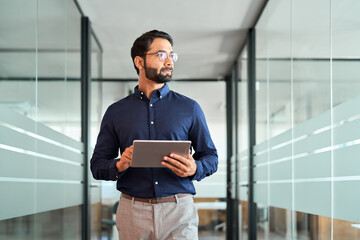 Busy Indian business man project manager looking away holding digital tablet walking in office. Professional businessman entrepreneur going in hallway holding tab working using tab computer.