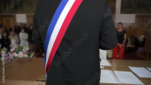A mayor from behind with a French tricolor scarf making a speech to celebrate a heterosexual marriage in a town hall. Blurred background on the bride and groom. French wedding with two happy lovers. photo