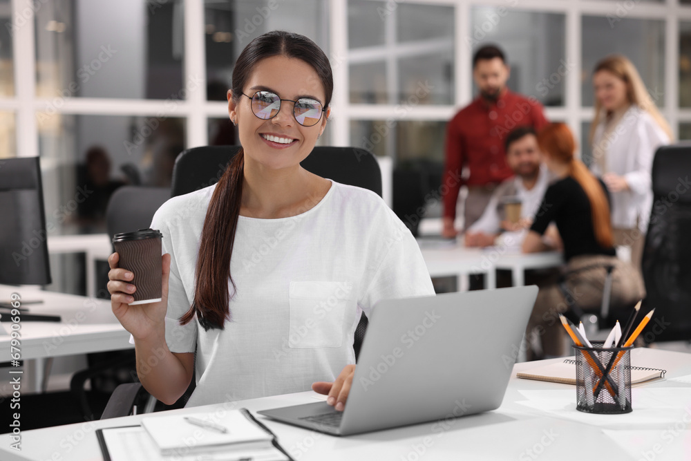 Team of employees working together in office. Happy woman with laptop and paper cup at table indoors Team of employees working together in office. Happy woman with laptop and paper cup at table indoors