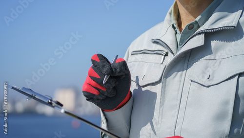 クリップボードを持つ作業服の男性・海と工業地帯│建設業・製造業・プラント イメージ