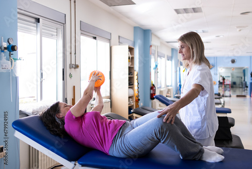 A middle aged woman is doing rehabilitation on a hospital bed while her physiotherapist is behind her teaching her some exercises.