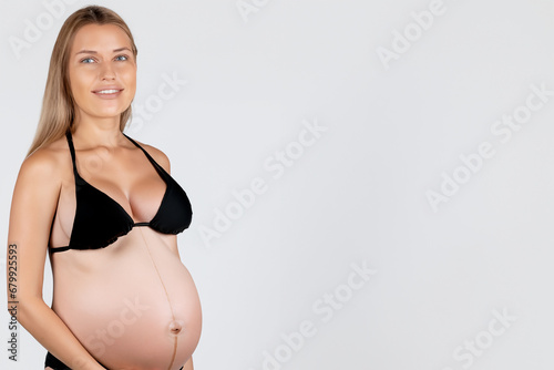 Portrey of a smiling blonde and blue eyed woman in an advanced stage of pregnancy in a black bra on a white background photo