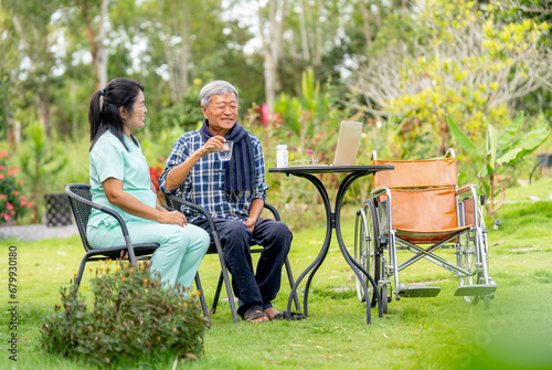 Asian senior man hold a glass of water and talk to nurse after take the medicine from her and they sit outdoor in the garden with wheelchair is set beside.