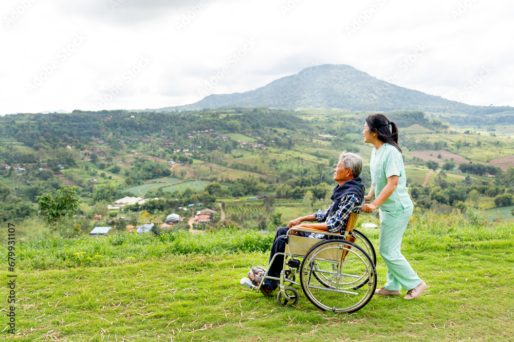 Asian nurse stand and hold wheelchair hand that sit by senior man and both stay near cliff with village and mountain in the background.