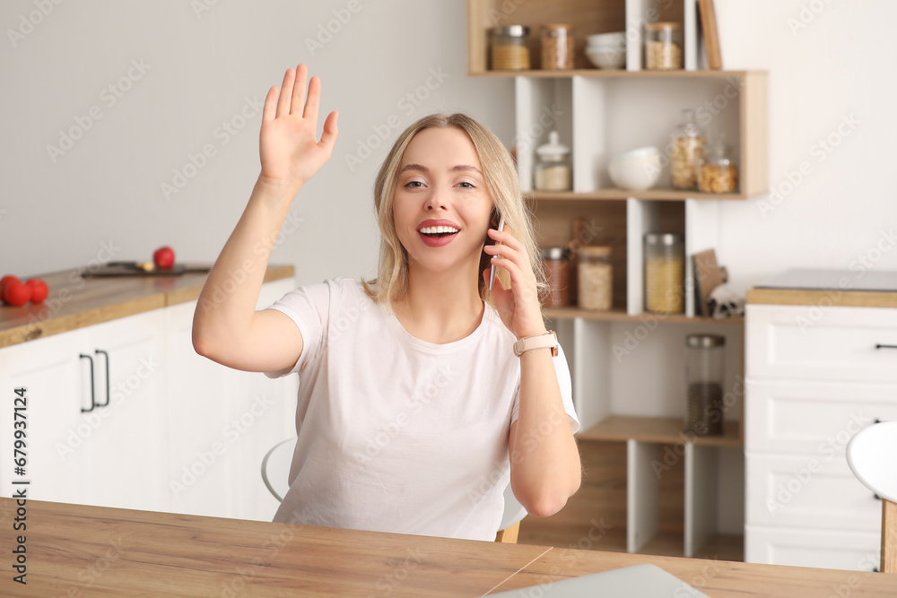Beautiful young woman talking by mobile phone and waving hand in kitchen at home Beautiful young woman talking by mobile phone and waving hand in kitchen at home