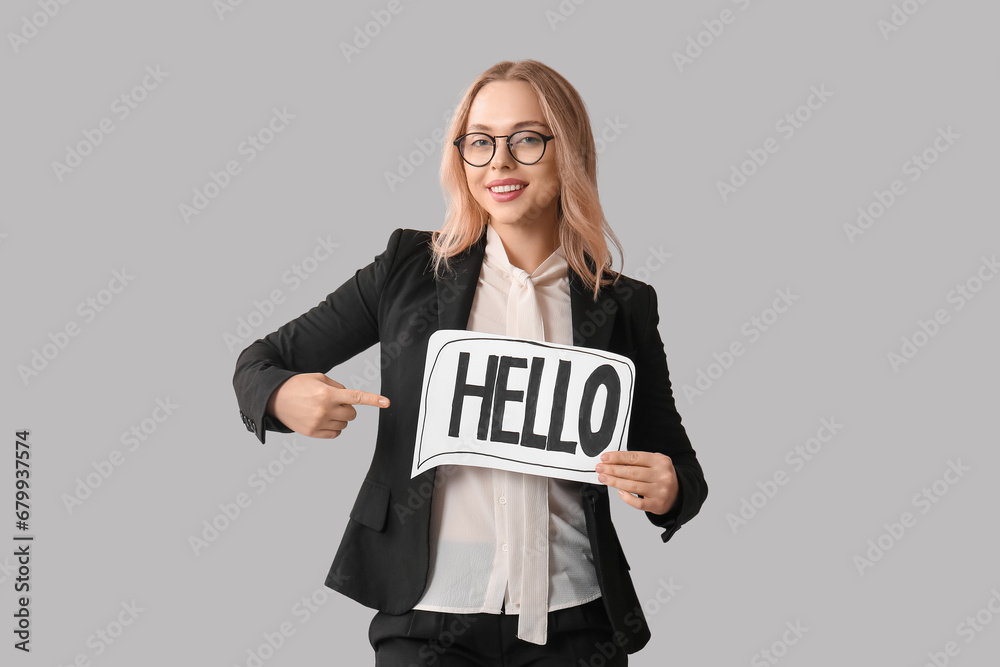 Beautiful businesswoman pointing at paper speech bubble with word HELLO on grey background Beautiful businesswoman pointing at paper speech bubble with word HELLO on grey background