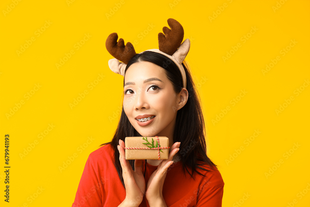 Young Asian woman in reindeer horns with Christmas gift box on yellow background Young Asian woman in reindeer horns with Christmas gift box on yellow background