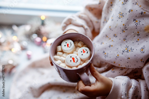 Unrecognizable little child girl holding cup with hot chocolate with marshmallows as snowman. Kid sitting near Christmas decorated window with lights. © Irina Schmidt
