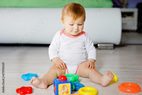 Adorable baby girl playing with educational toys in nursery. Happy healthy child having fun with colorful different toys at home. Kid trying to build plastic pyramid and using blocks with letters