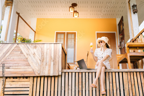 Fototapeta Naklejka Na Ścianę i Meble -  Asian female tourist Come relax on holiday. At a beautiful resort made of wood Sitting and relaxing in the front of the room, holding a wine glass, laptop, wearing a dress, hat, and glasses.