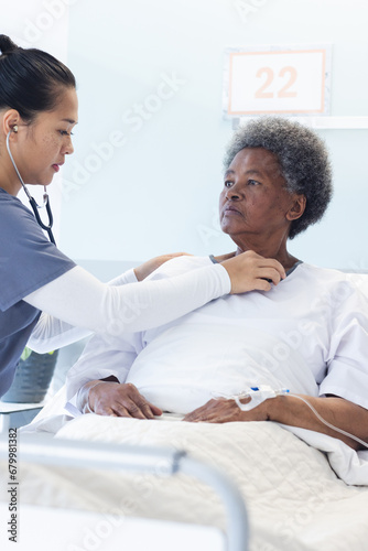 Diverse female doctor testing senior female patient using stethoscope in hospital room