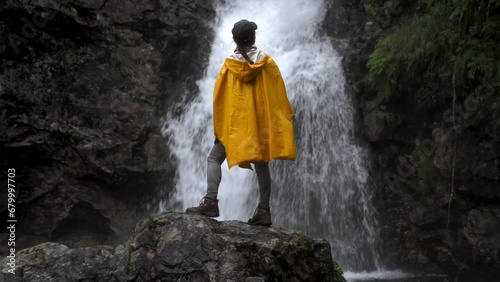 backview of a full lenght silhouette of an asian woman in yellow hooded raincoat and black cap with waterfall on the background photo