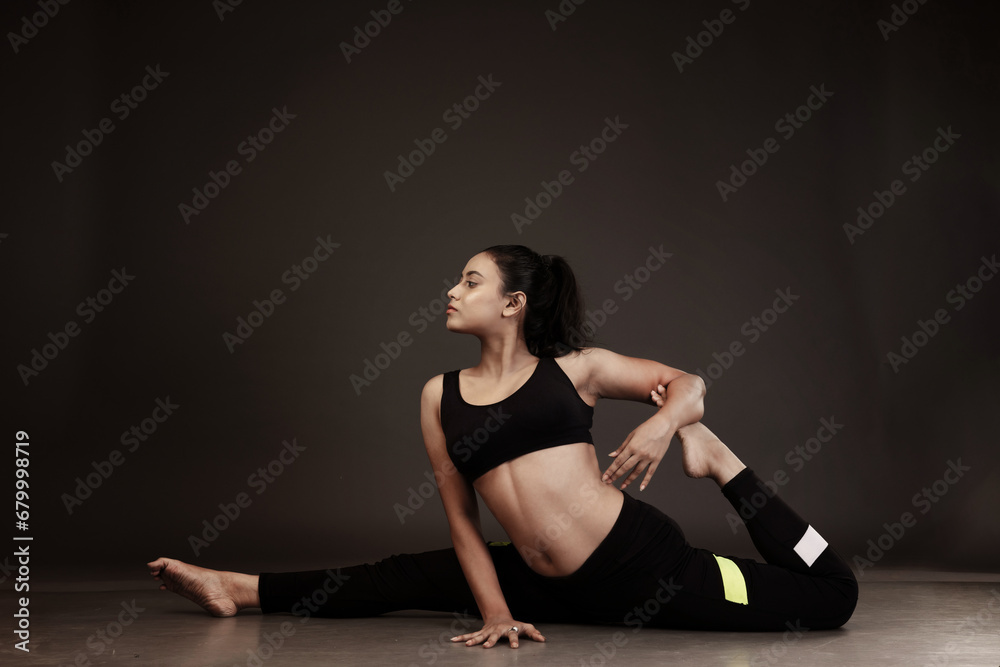 Obraz premium Slim girl performing yoga arts wearing black sports dress in black background performing yoga poses with a flexible body