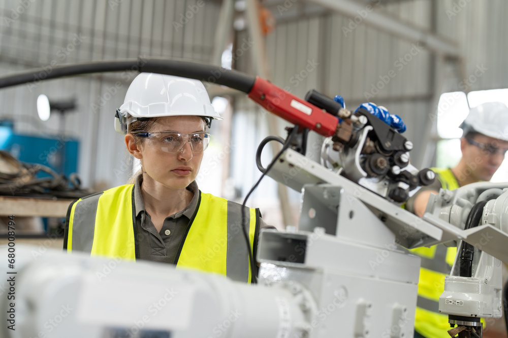 Female automation engineer in safety vest with white helmet checking and inspecting new welding robot arm machine in an industrial factory