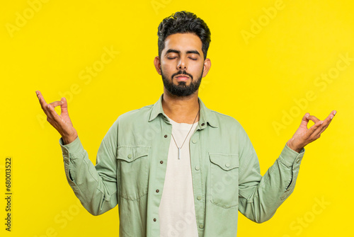 Keep calm down, relax, rest. Concentrated happy Indian man meditating breathes deeply with mudra yoga gesture, eyes closed, peaceful mind, taking a break. Arabian Hindu guy on yellow studio background