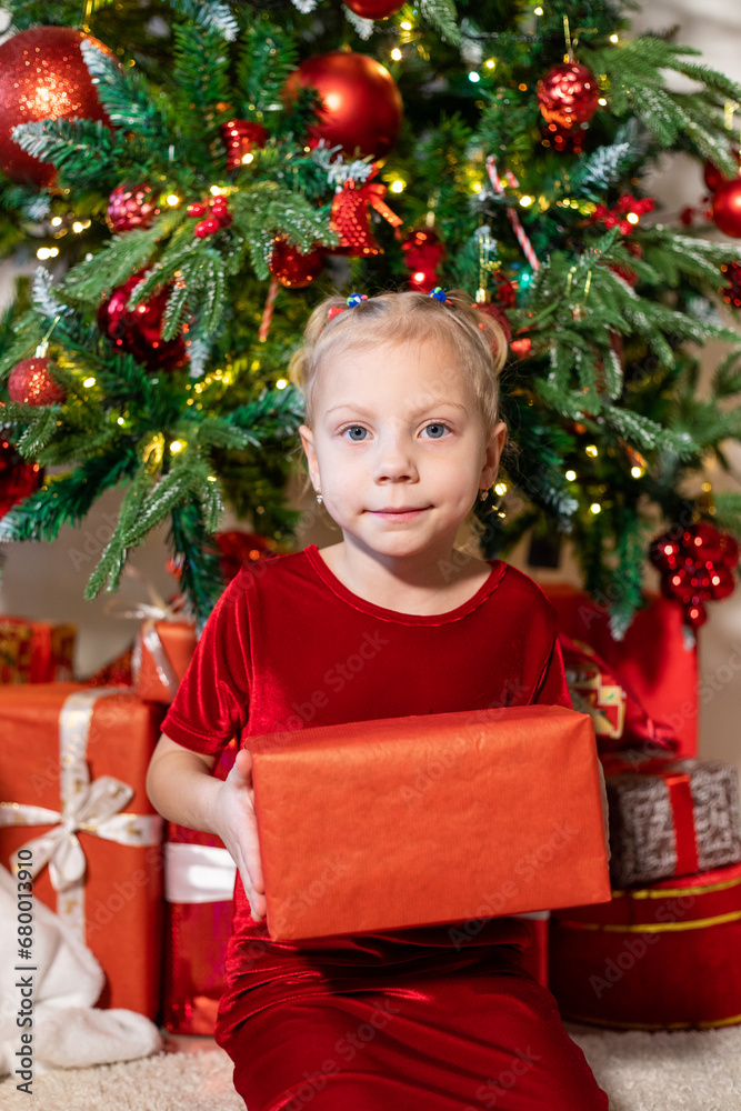 Cute girl with a gift in her hands sits near the Christmas tree.