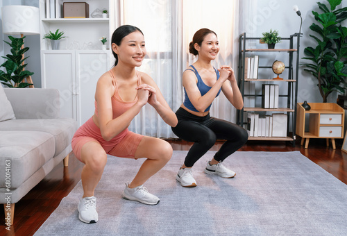 Vigorous energetic woman with trainer or workout buddy doing exercise at home. Young athletic asian woman strength and endurance training session as home workout routine with squat.