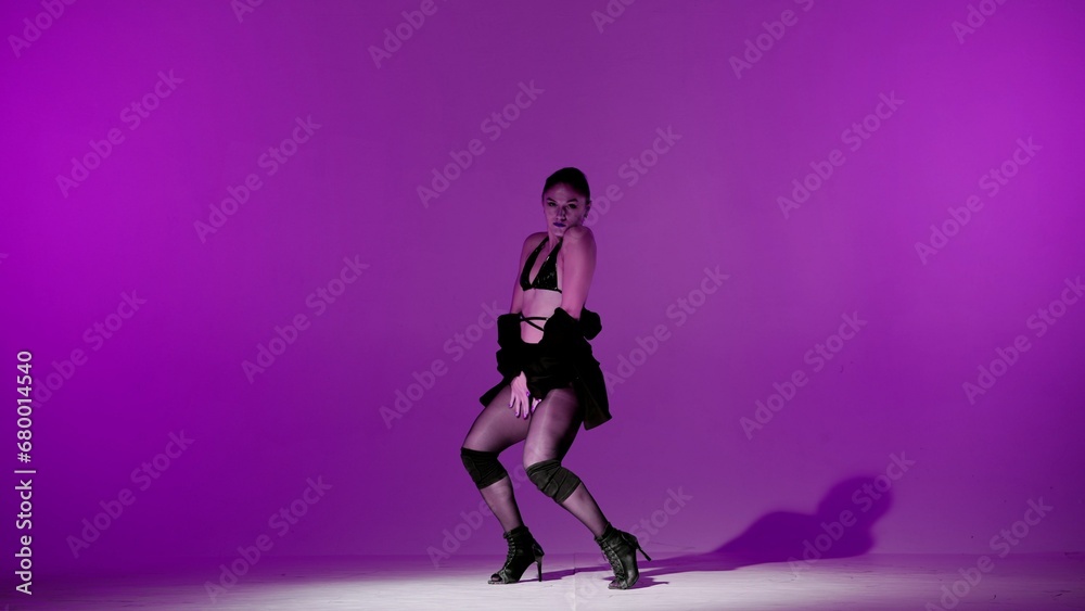 On purple background. Young woman, showing dance moves towards high heels. A spotlight beam shines on her and gives her a shadow, the edges of the background are darkened. She is sexy, rhythmic