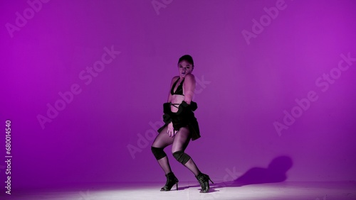 On purple background. Young woman, showing dance moves towards high heels. A spotlight beam shines on her and gives her a shadow, the edges of the background are darkened. She is sexy, rhythmic