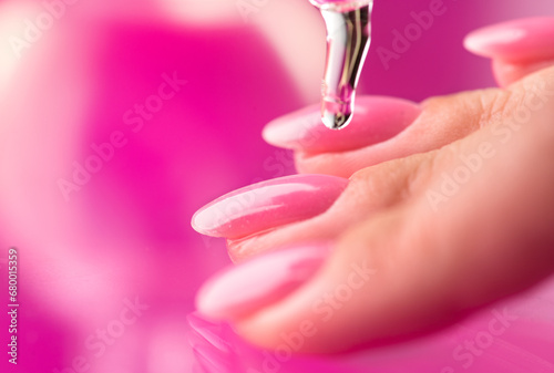 Applying cuticle oil in pipette, on fingers, manicure salon. Nail care, polish, pink shellac UV gel, varnish, manicure process in beauty salon. Over pink background. Application of nails oil closeup