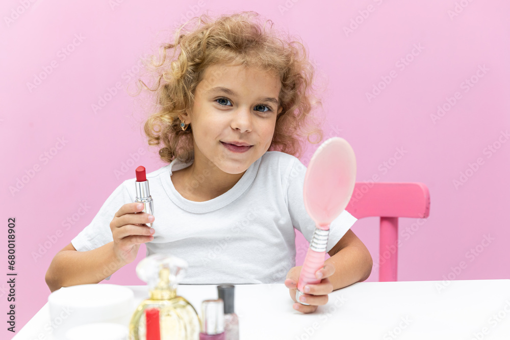 Happy little girl with blonde curly hair puts lipstick on her lips looking in the mirror at home on pink background. Little trendy. little girl applying makeup. High quality photo with copy space Happy little girl with blonde curly hair puts lipstick on her lips looking in the mirror at home on pink background. Little trendy. little girl applying makeup. High quality photo with copy space