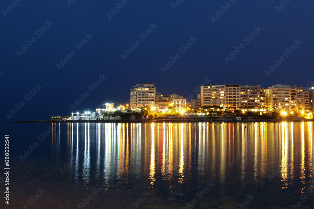 Long reflections of seaside resort night lights on calm water of Mediterranean sea bay.