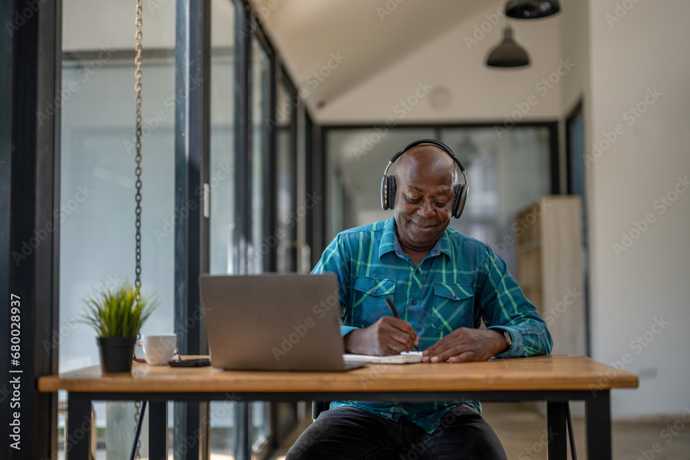 Fototapeta premium Senior black man wearing headphones enjoying listening to music or something interesting to relax while working. Study and take notes of interesting and useful things.