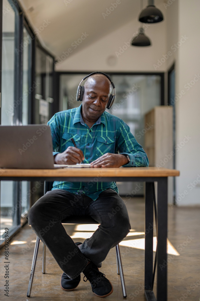 Full size portrait of senior black man wearing headphones enjoying listening to music or something interesting to relax while working. Study and take notes of interesting and useful things.
