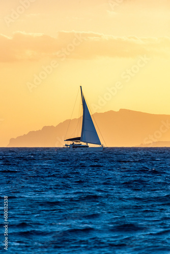 Sailboat sailing at deep blue sea at sunset with background of vivid orange sky