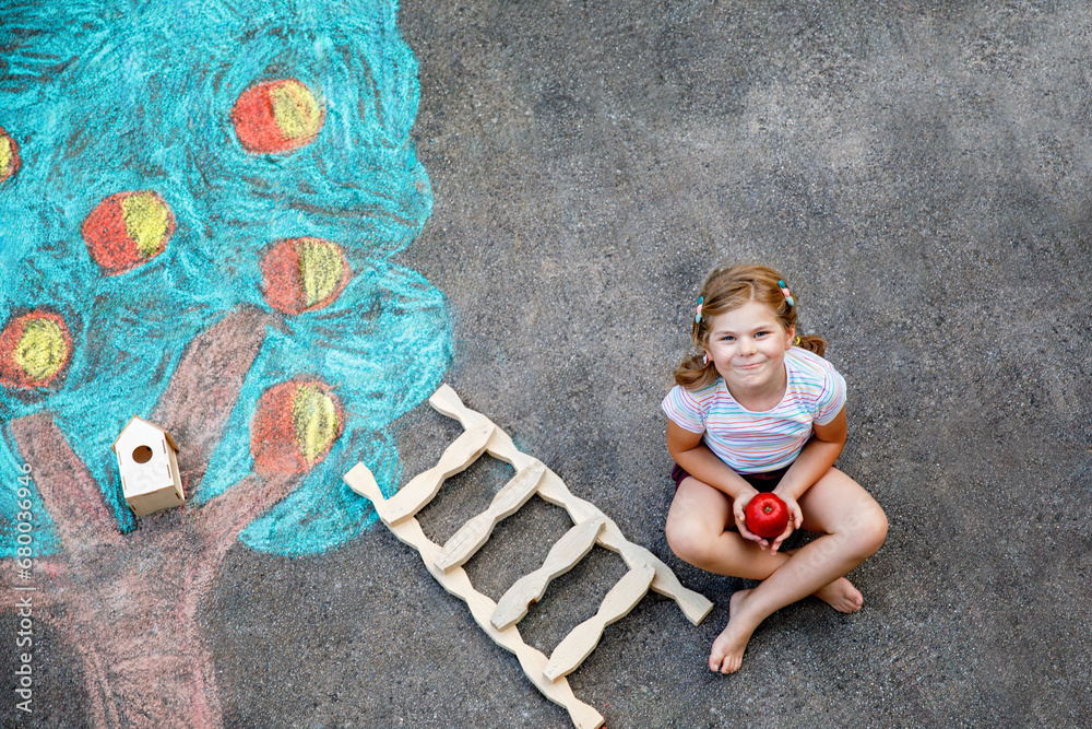 Naklejka premium Cute little girl painting with colorful chalks apples harvest from apple tree on asphalt. Cute preschool child with having fun with chalk picture. Creative leisure for children, drawing and painting.
