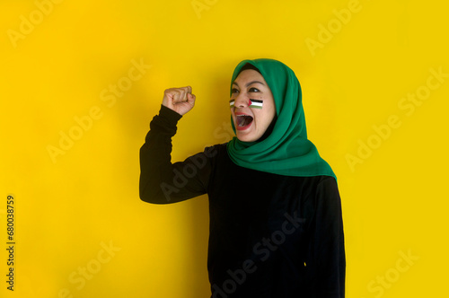 Young muslim woman wearing hijab with Palestine flag sticker on her cheek raising fist and screaming aloud 
