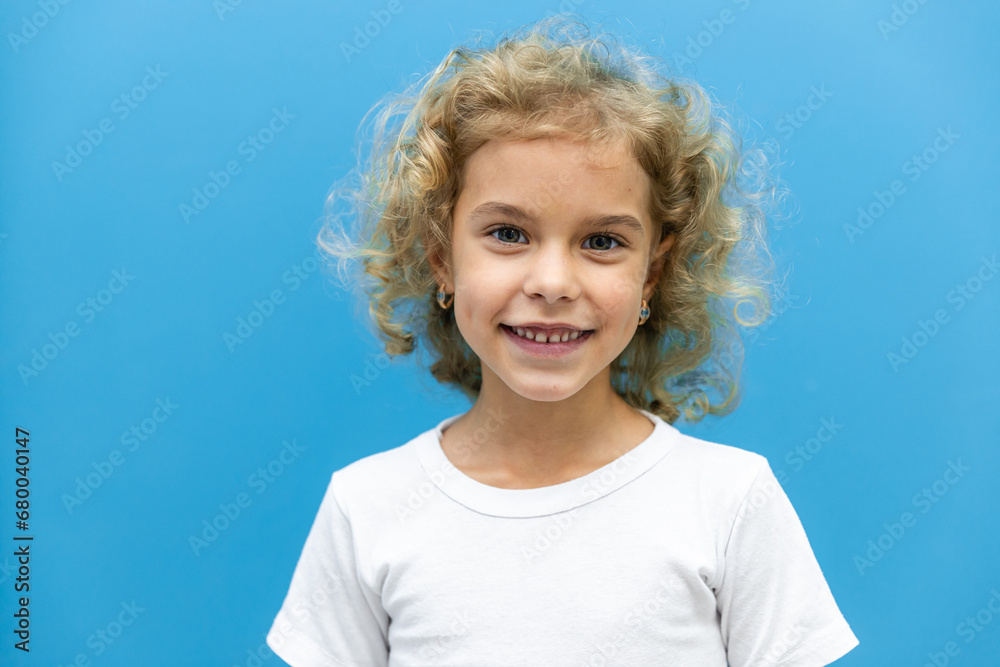 Close up head shot portrait with smiling little blonde curly hair girl. Concept happy and beauty kid with good healthy teeth for dental on blue background, six year child looking at camera and posing - obrazy, fototapety, plakaty 