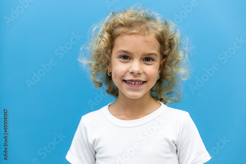 Close up head shot portrait with smiling little blonde curly hair girl. Concept happy and beauty kid with good healthy teeth for dental on blue background, six year child looking at camera and posing