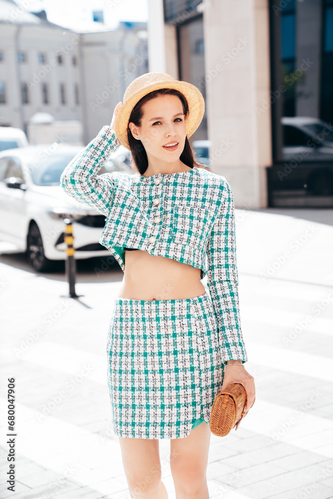Young beautiful smiling woman in trendy summer green jacket and skirt clothes. Sexy carefree model posing in the street at sunset. Positive brunette female. Cheerful and happy. In hat, holds clutch