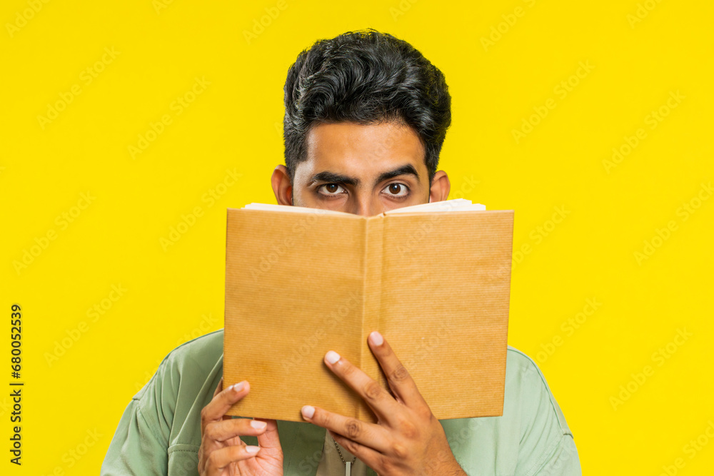 Sneaky cunning relaxed Indian young man reading interesting fairytale story book, leisure hobby, knowledge wisdom, education, learning, study, wow. Arabian Hindu guy isolated on yellow background - obrazy, fototapety, plakaty 