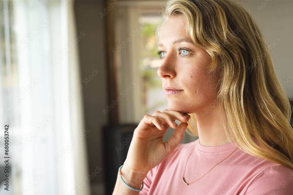 Thoughtful blonde caucasian woman looking out window in sunny living room at home Thoughtful blonde caucasian woman looking out window in sunny living room at home