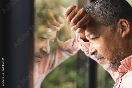 Thoughtful senior biracial man wearing checked shirt and leaning on window at home photo