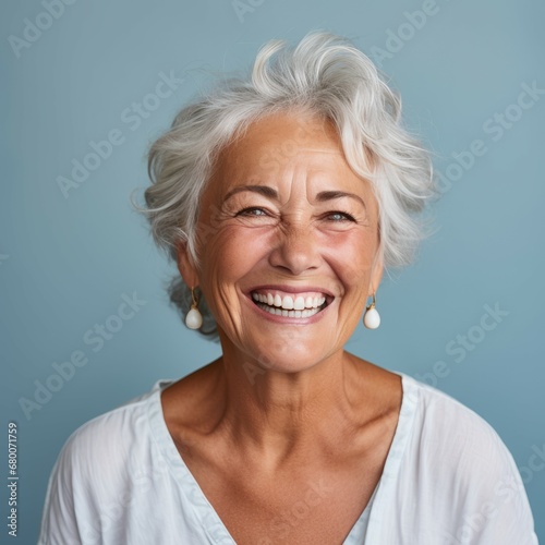 Portrait of a smiling senior woman with wavy gray hair on a blue background. Happy old Caucasian woman with a smile in a blue shirt looking at camera. Cheerful European woman with shiny white teeth.