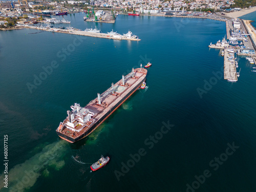 A massive cargo ship bulk carrier leaves the seaport, accompanied by tugboats, aerial view