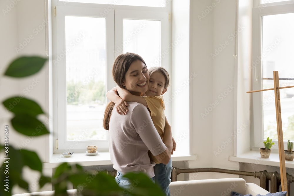 Caring young mother embracing tightly little daughter with eyes closed, shows love and affection, enjoy tender moment. Loving family spend weekend time together at home. Happy motherhood, childhood