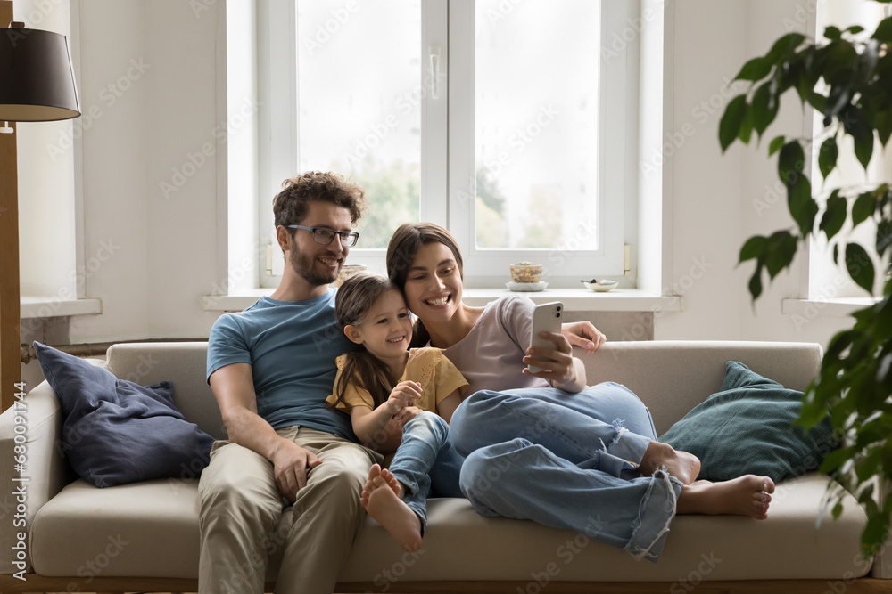 Smiling young spouses sit on sofa with little daughter hold smartphone, take self-portrait on camera. Family spend weekend time make video conference, using internet and videocall app on mobile phone Smiling young spouses sit on sofa with little daughter hold smartphone, take self-portrait on camera. Family spend weekend time make video conference, using internet and videocall app on mobile phone