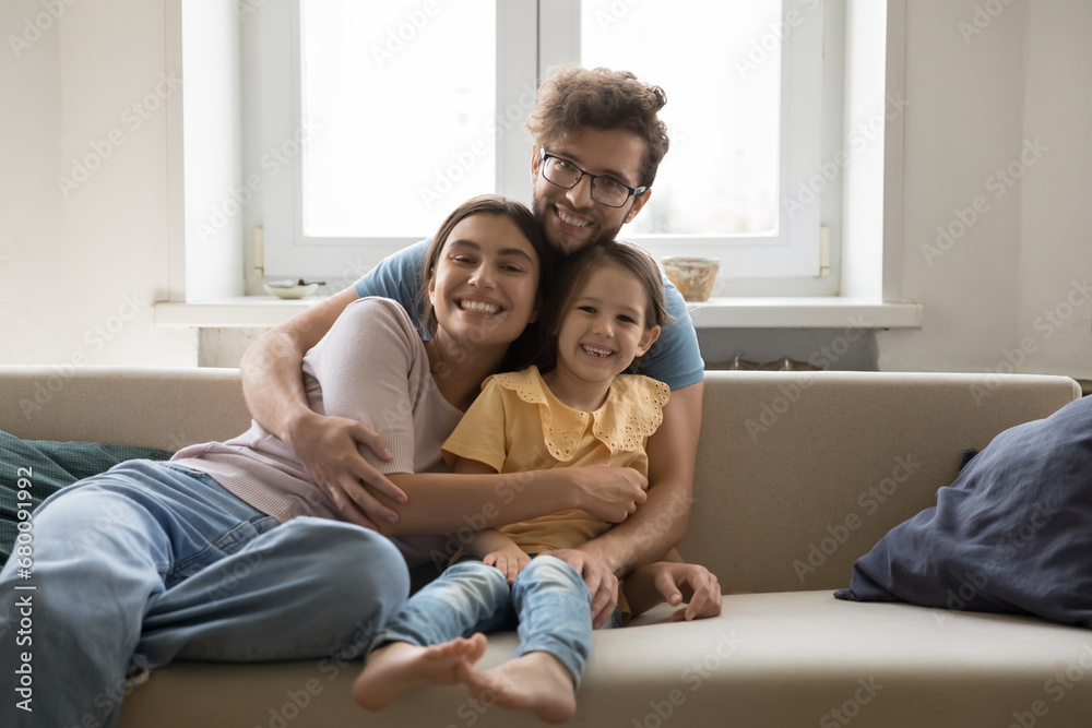 Loving young man hug wife and little daughter sitting on sofa, happy family smile pose looking at camera. Happy parents spend weekend time with cute kid at home, enjoy time together. Parenthood, love Loving young man hug wife and little daughter sitting on sofa, happy family smile pose looking at camera. Happy parents spend weekend time with cute kid at home, enjoy time together. Parenthood, love