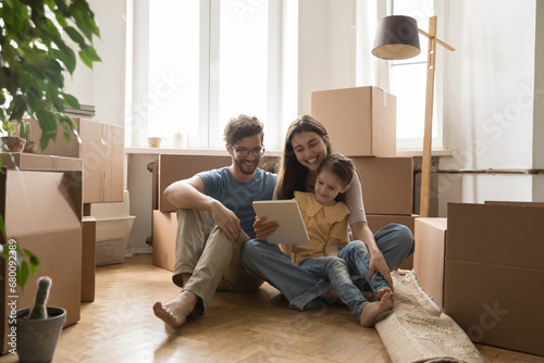 Young family with child sit on floor at new flat near boxes with stuff use tablet, browse internet, discuss furniture purchase. Spouses buy items for new house on relocation. Move-in day, e-commerce © fizkes