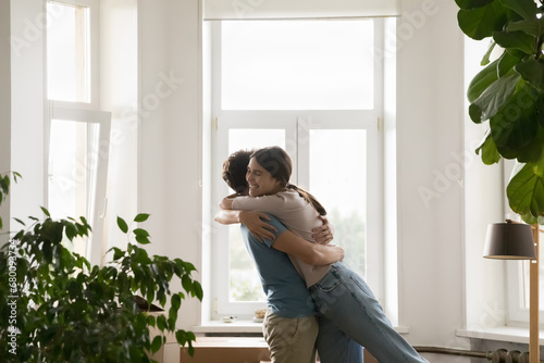 Overjoyed loving young spouses cuddling standing in living room with cardboard boxes, congratulate each other with first dwelling purchase, celebrating relocation day, swirling, hugging feel happy