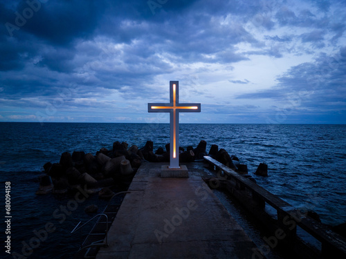 A large Christian cross stands at the edge of a pier against a dramatic sky and sea, seen from above