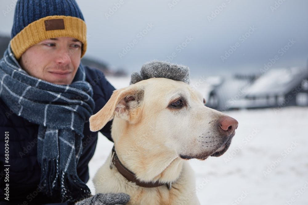 Man dressed in warm jacket, with scarf and cap stroking his dog. Pet owner and labrador retriever against mountain village in winter. Selective focus on dog.. Man dressed in warm jacket, with scarf and cap stroking his dog. Pet owner and labrador retriever against mountain village in winter. Selective focus on dog..