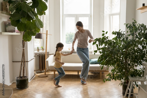 Beautiful mother teach cute little kid to dance, dancing to favorite music in living room, enjoy active weekend at home. Daughter and mom holding hands, moving in cozy flat. Motherhood, hobby, fun photo
