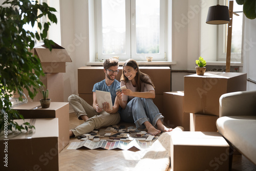 Fotografia On relocation day happy young wife and husband sit on floor giving high five, boxes with belongings and heap of color samples tiles, renovation brochures nearby