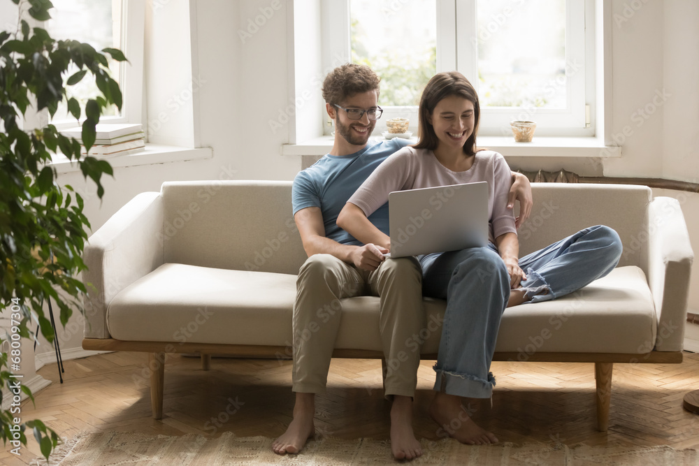 Young pretty happy couple sit on cozy sofa in living room with laptop on laps looking at screen, enjoy movie or funny videos online. Smiling family e-shopping, spend weekend time at home on internet